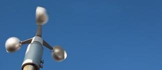 A three-cup anemometer with blurred cups spins against a bright blue sky, measuring wind speed.