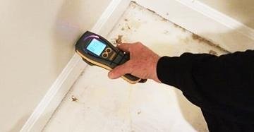 A hand holds a digital moisture meter against a wall, scanning for hidden moisture above a stained, damp-looking floor.