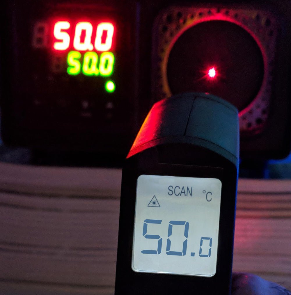 A hand holds an infrared thermometer displaying "SCAN 50.0 °C", aimed with a red laser dot at a circular object. In the background, a digital display unit shows "50.0" in red and "50.0" in green.