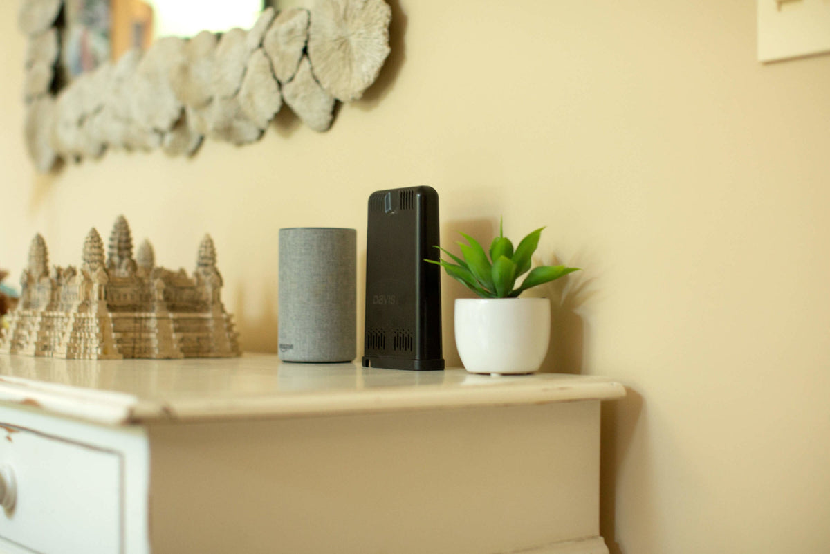 Data collection hub by Davis Instruments with ventilation slots, sitting next to a gray cylindrical speaker and small potted plant.