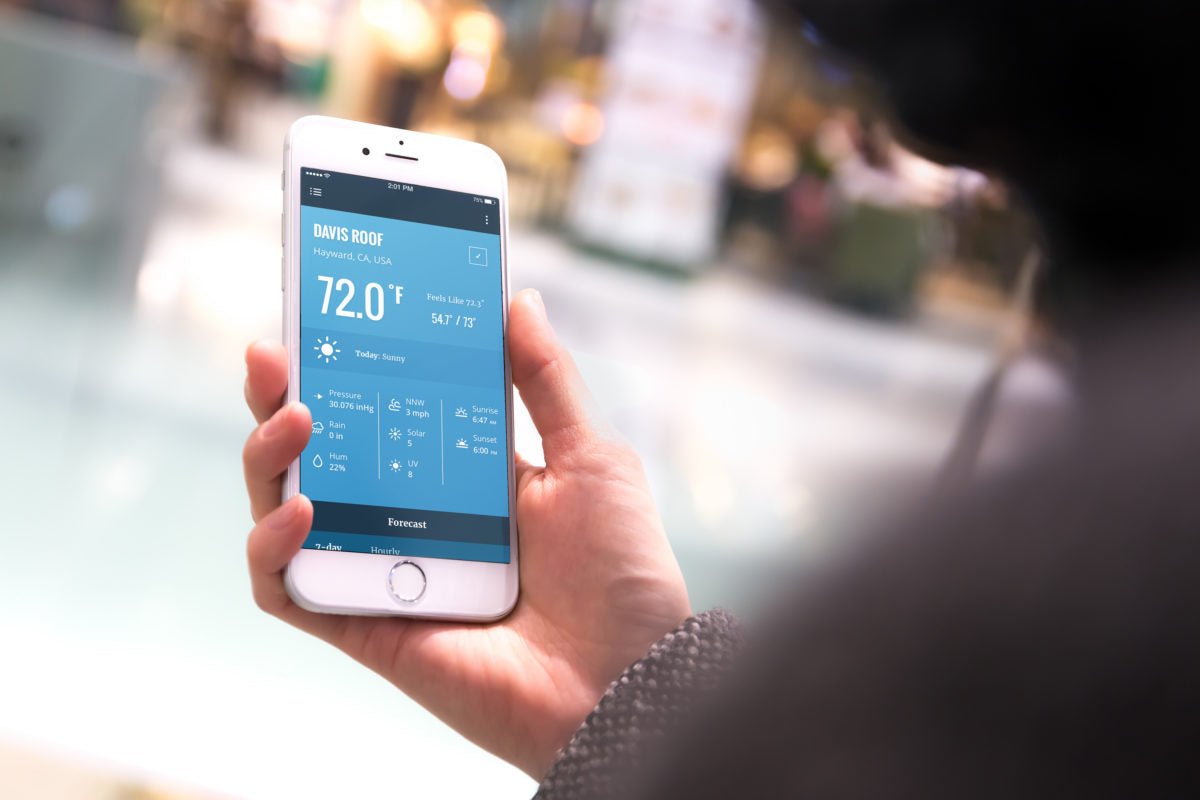 Weather app interface by Davis Instruments showing a sunny forecast on a smartphone held in hand.